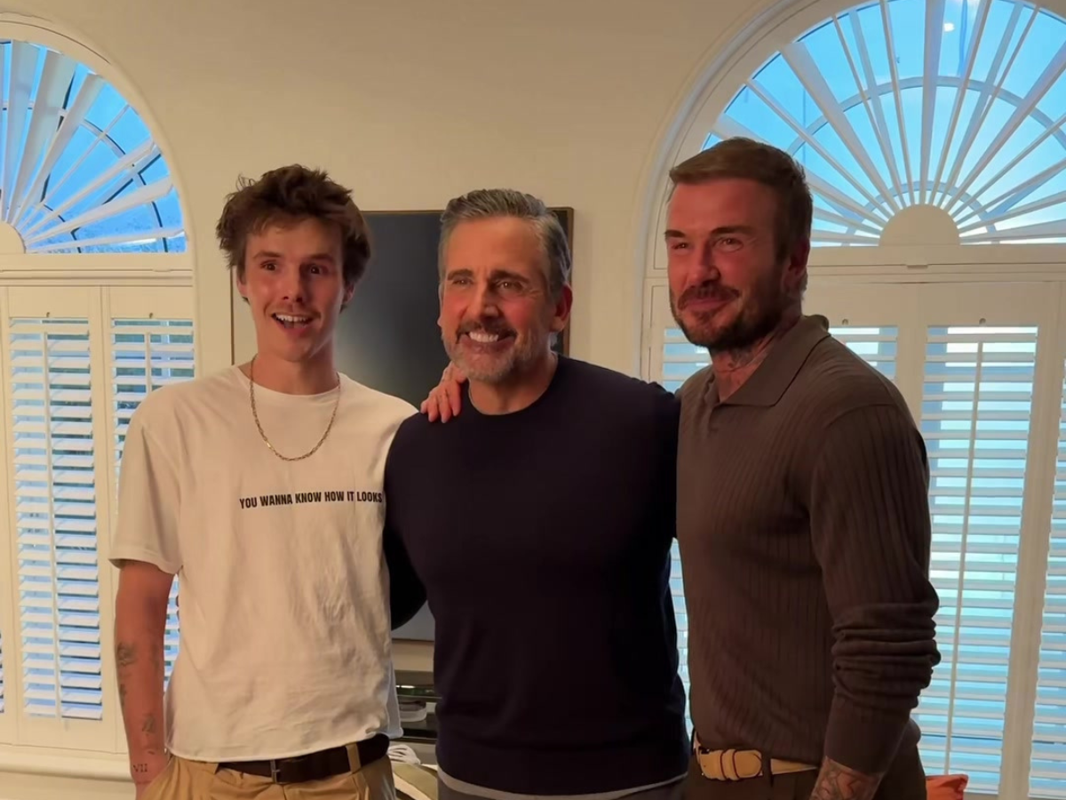 Three men standing together indoors, smiling, with arched windows and blinds behind them.