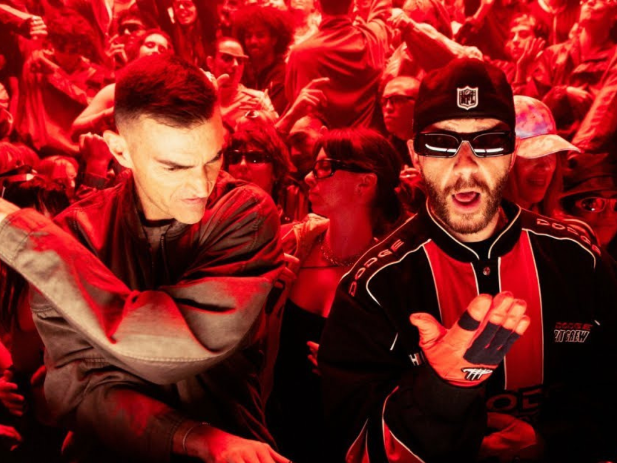 Two men in a packed crowd: left is in a brown jacket looking down, right wears a red-and-black racing suit with gloves and a cap, surrounded by fans.