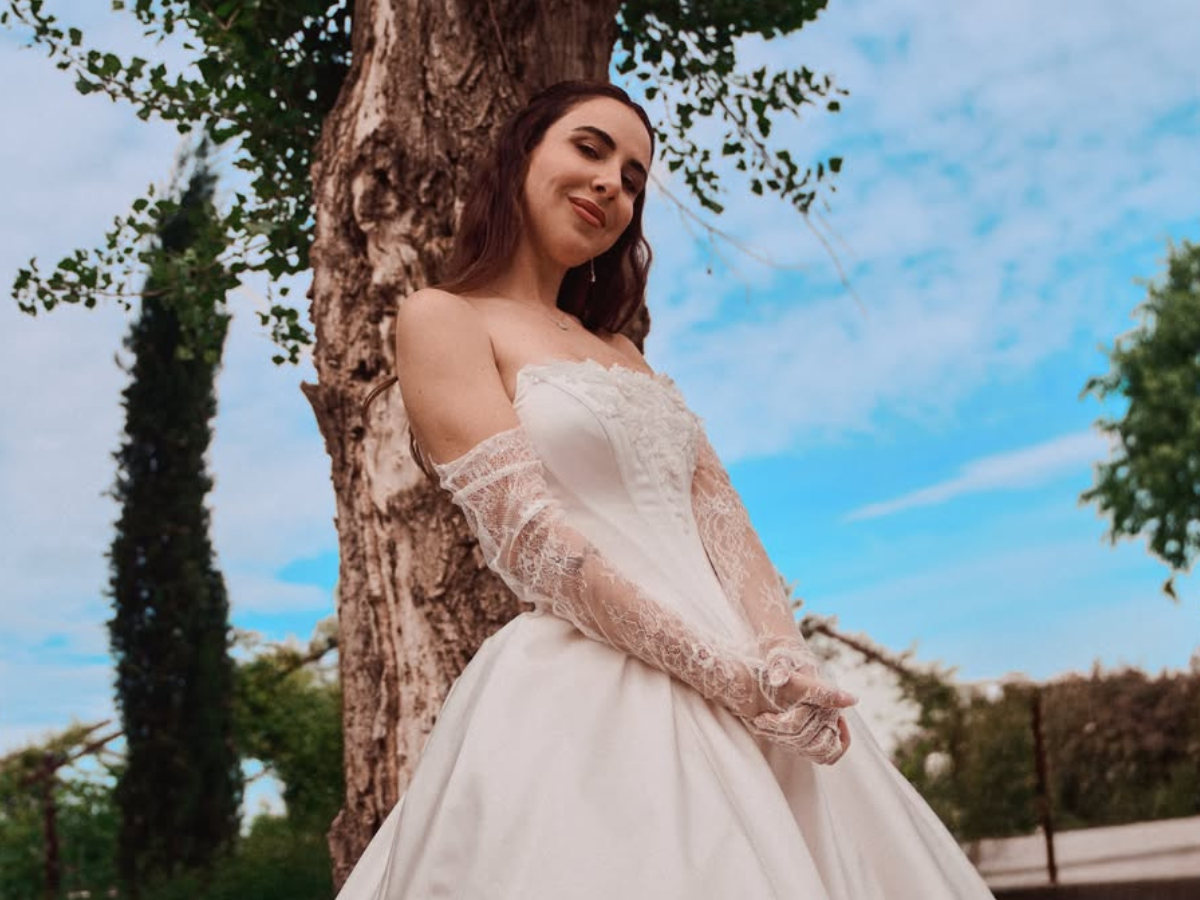 Bride in a white wedding dress with lace sleeves, standing beside a tree outdoors with a bright blue sky in the background.