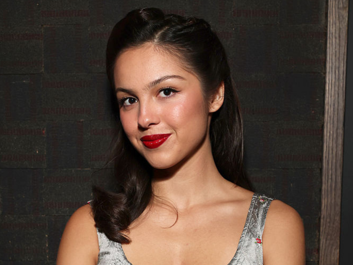 Portrait of a woman with dark hair, bright red lipstick, and a silver dress, smiling softly against a dark background.