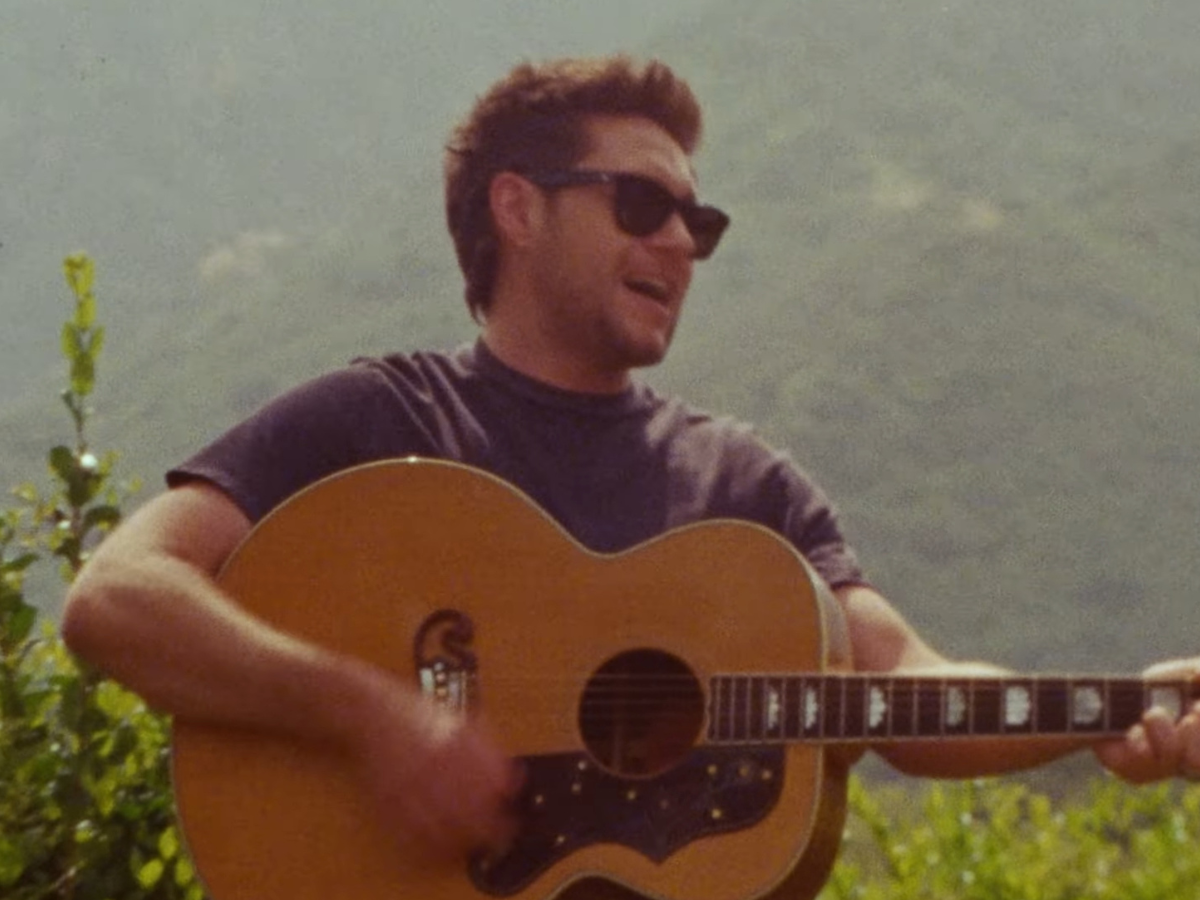 Man wearing a dark shirt and sunglasses playing an acoustic guitar outdoors in a sunny setting with hills in the background, mid-strum.