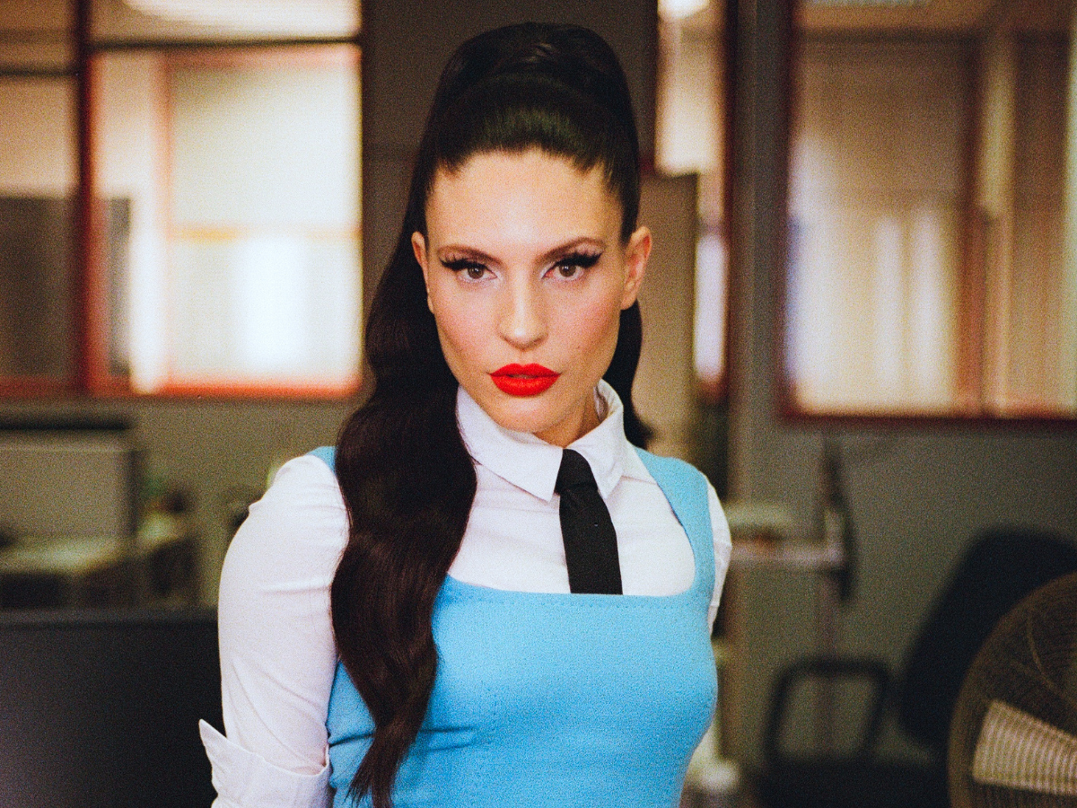 Portrait of a woman with retro hairstyle, wearing a light blue dress over a white blouse and black tie in an office setting.