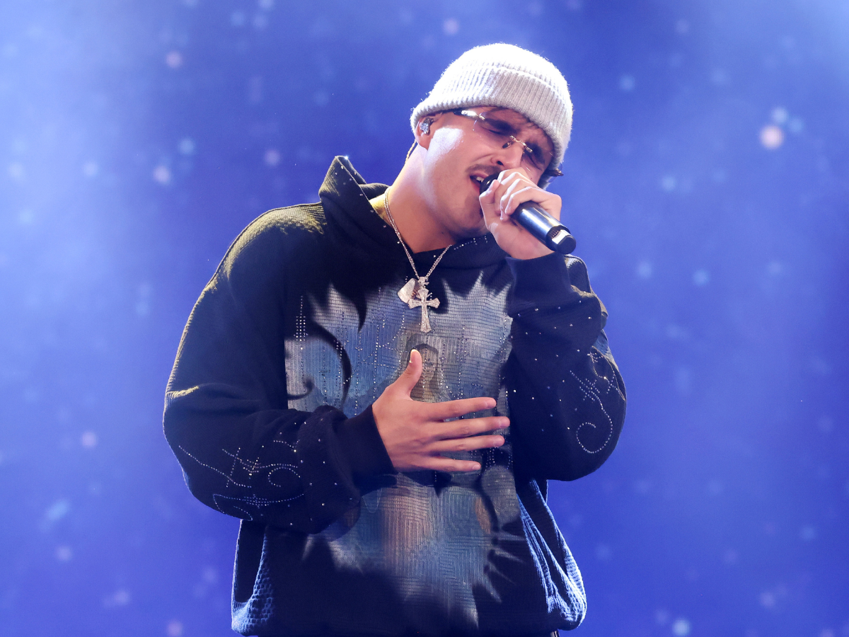 Male singer on stage wearing a light beanie, sunglasses, and a dark hoodie with a cross necklace, microphone in hand, performing passionately.