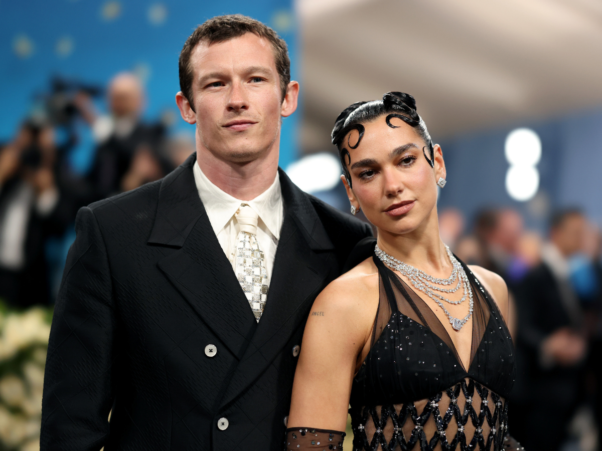 Man in a black double-breasted suit and cream tie posing with a woman in a black beaded dress on a red carpet at a formal event.