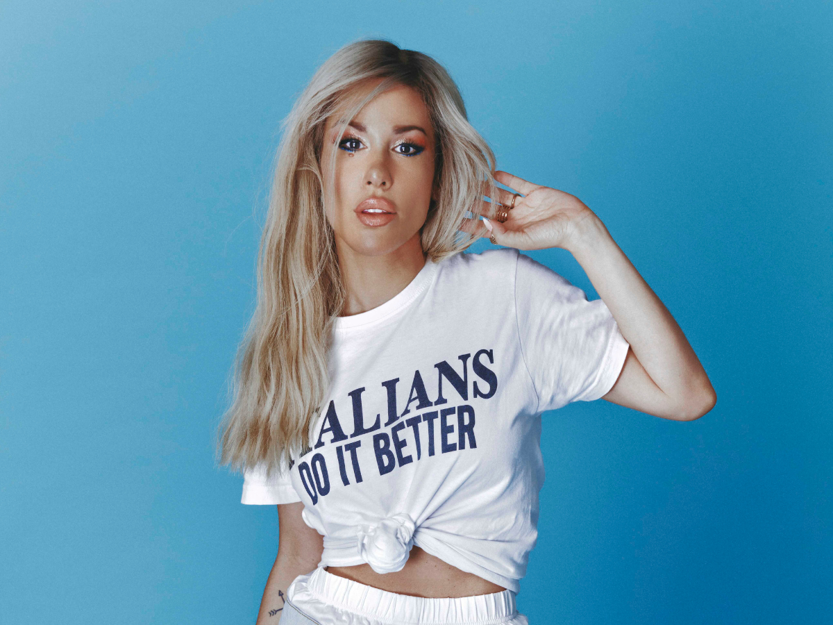 Portrait of a blonde woman posing against a blue background, wearing a knotted white T-shirt that reads 'ALLIANS DO IT BETTER'