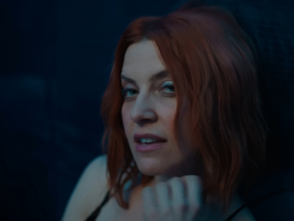 Close-up of a woman with red hair looking at the camera in dim blue lighting, hand near her chin.