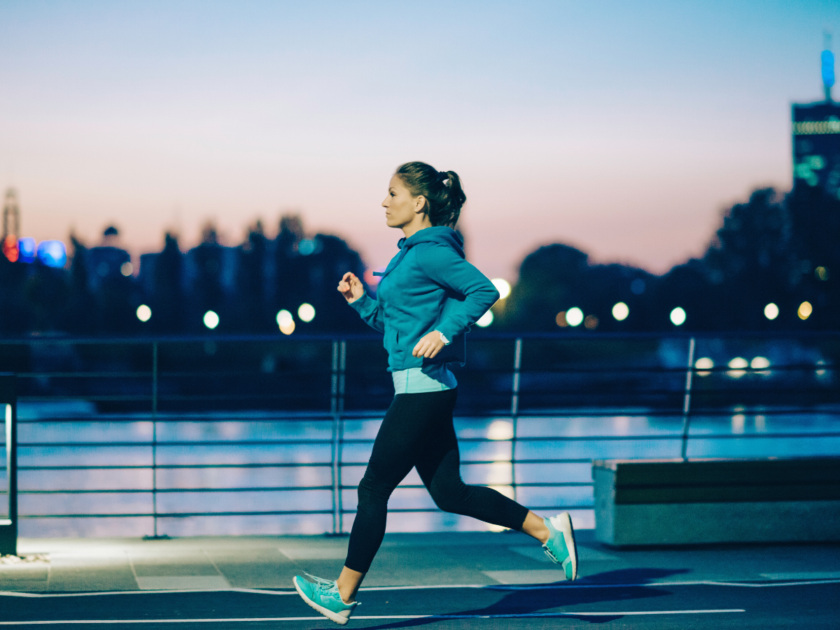 Woman running along a waterfront promenade at dusk, wearing a teal hoodie, black leggings, and teal running shoes with her hair in a ponytail.