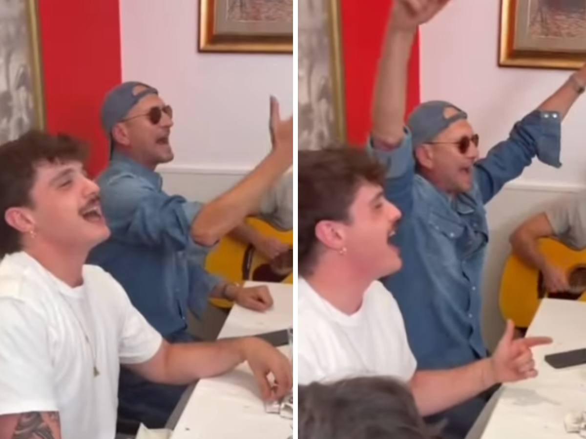 Two men sing and cheer at a table; a guitarist in a denim shirt and cap plays in the background.