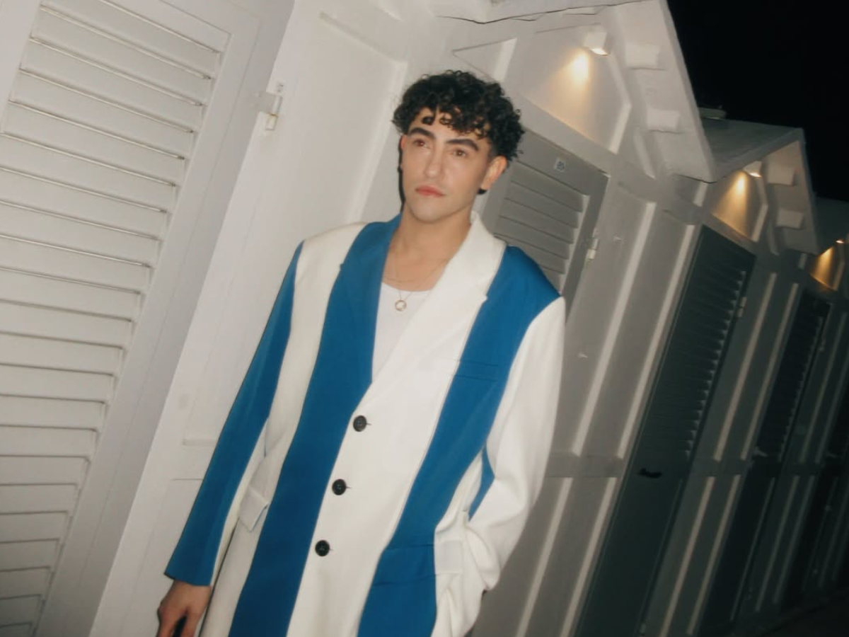 Young man with curly hair wearing a white and blue color-block blazer, standing in a white hallway at night.