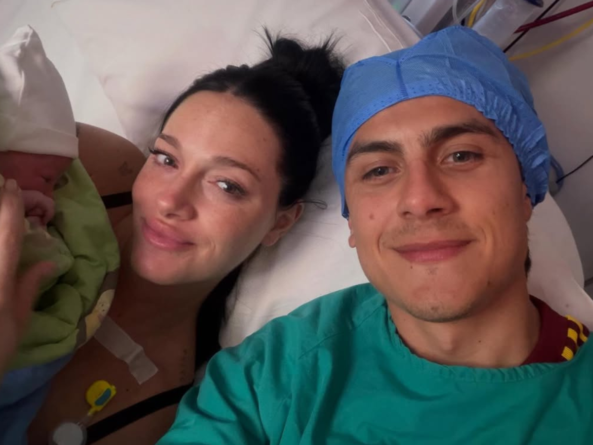 New parents in a hospital bed with their newborn baby; mother holds the baby, father wearing a blue surgical cap smiles nearby.