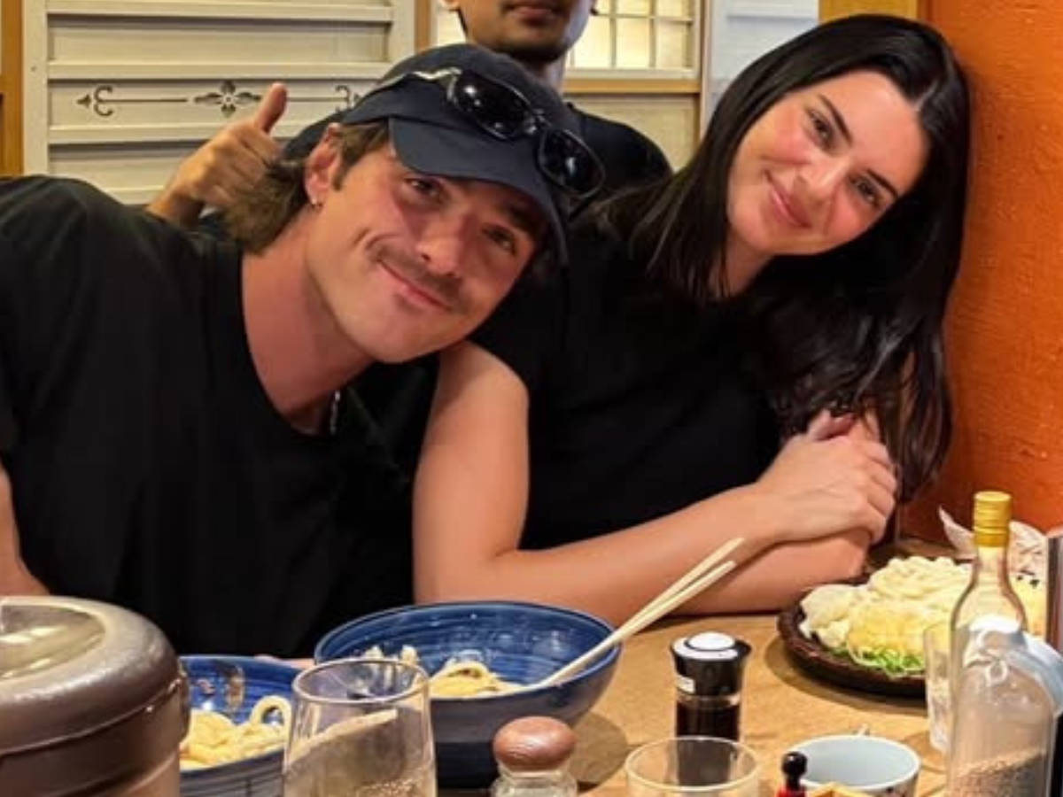 Two friends at a restaurant table, smiling at the camera; man wearing a cap with sunglasses on it and a woman with long dark hair beside him