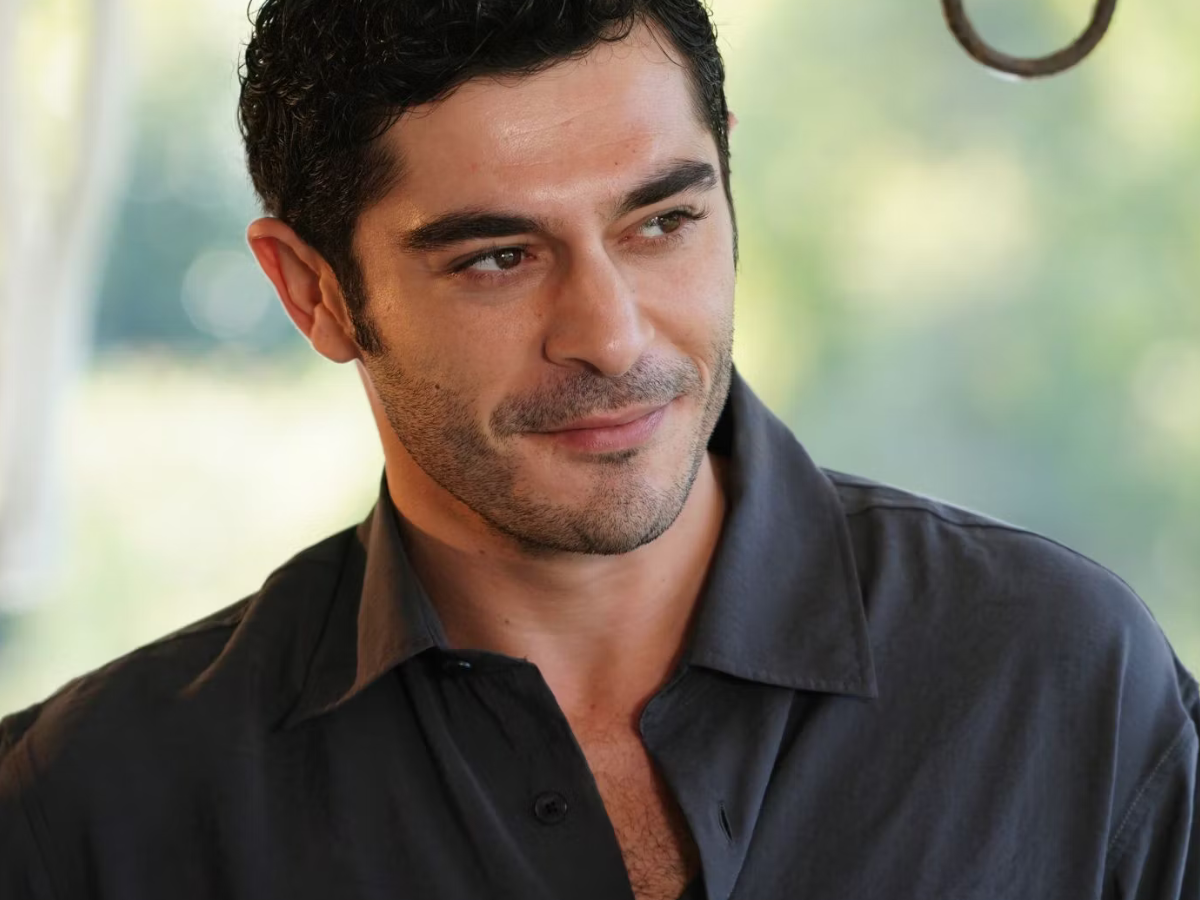 Close-up portrait of a man with dark hair and stubble, wearing a dark shirt, looking to the right with a soft expression.