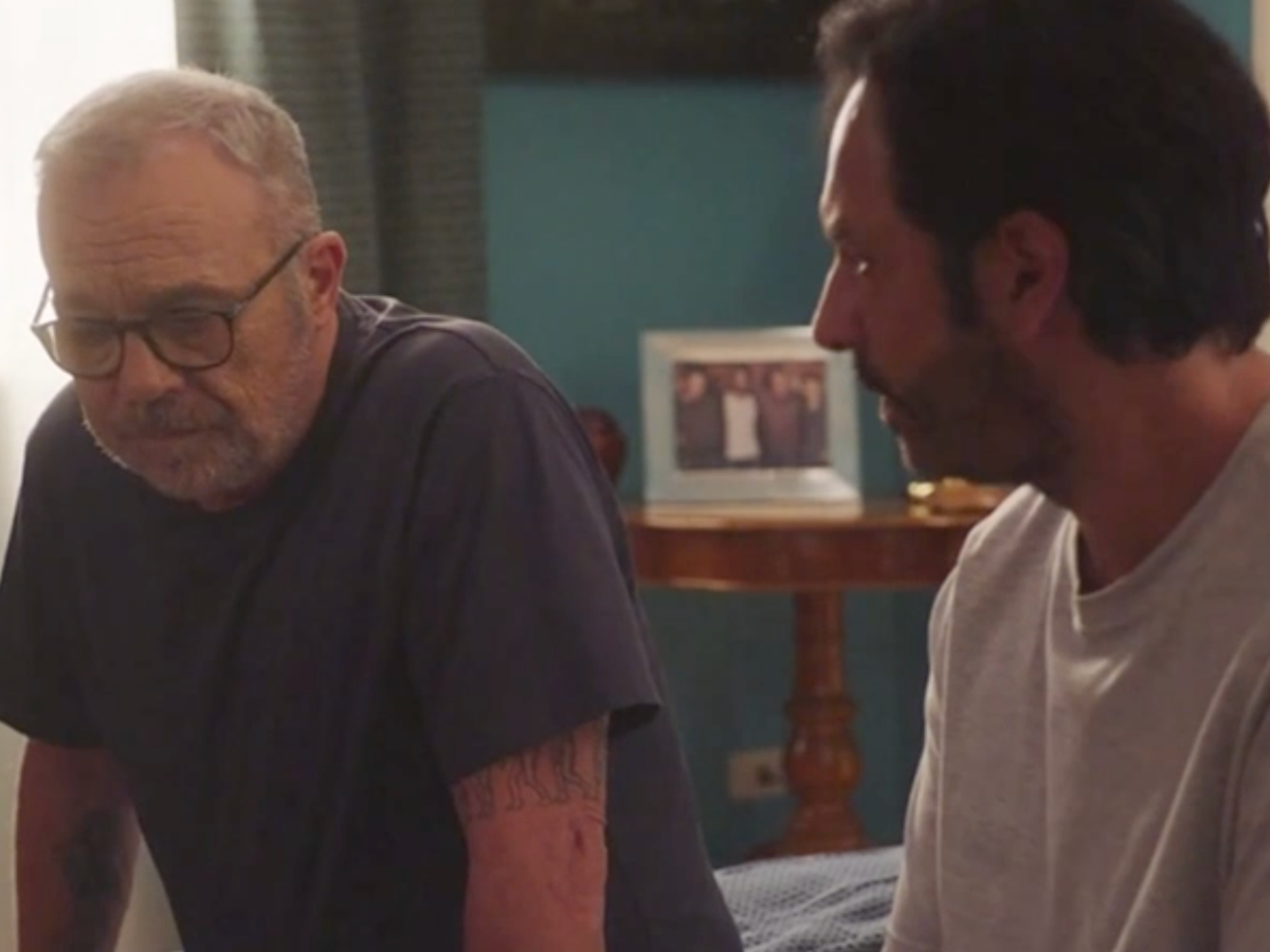 Two men sit indoors in conversation; older man with glasses and gray hair on the left, younger man on the right.