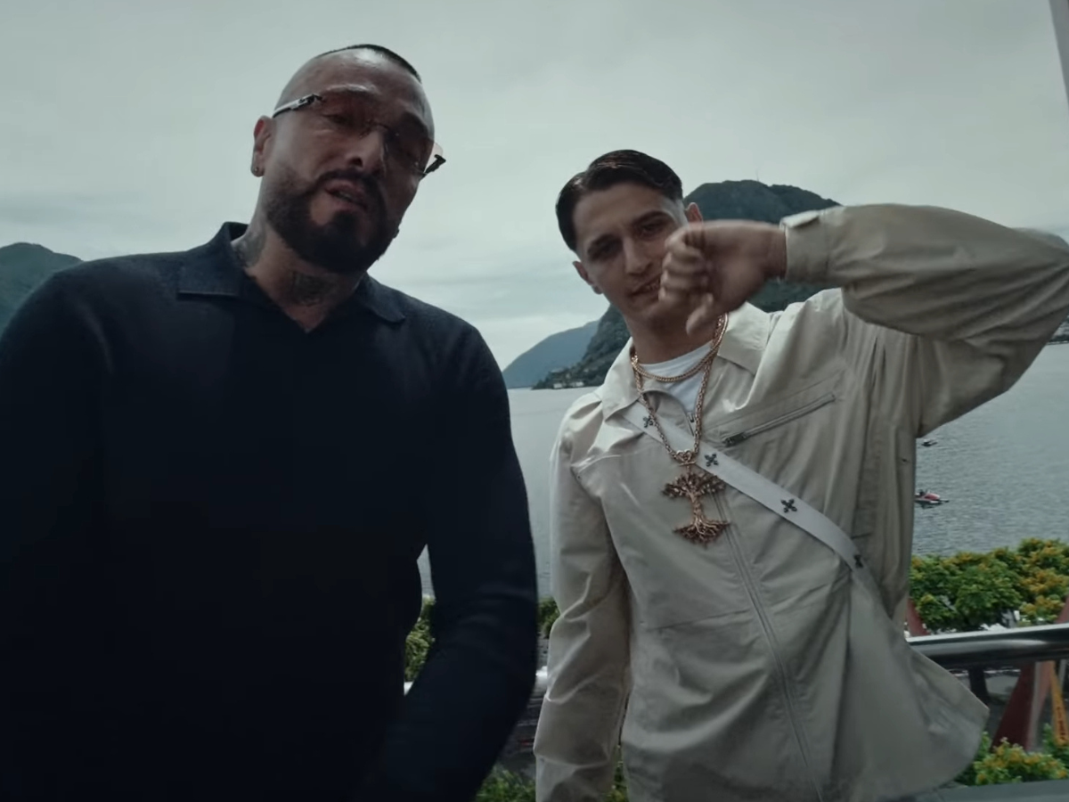 Two men pose for a photo on a terrace with a lake and mountains in the background, one in a black shirt and the other in a beige jacket showing a raised fist.