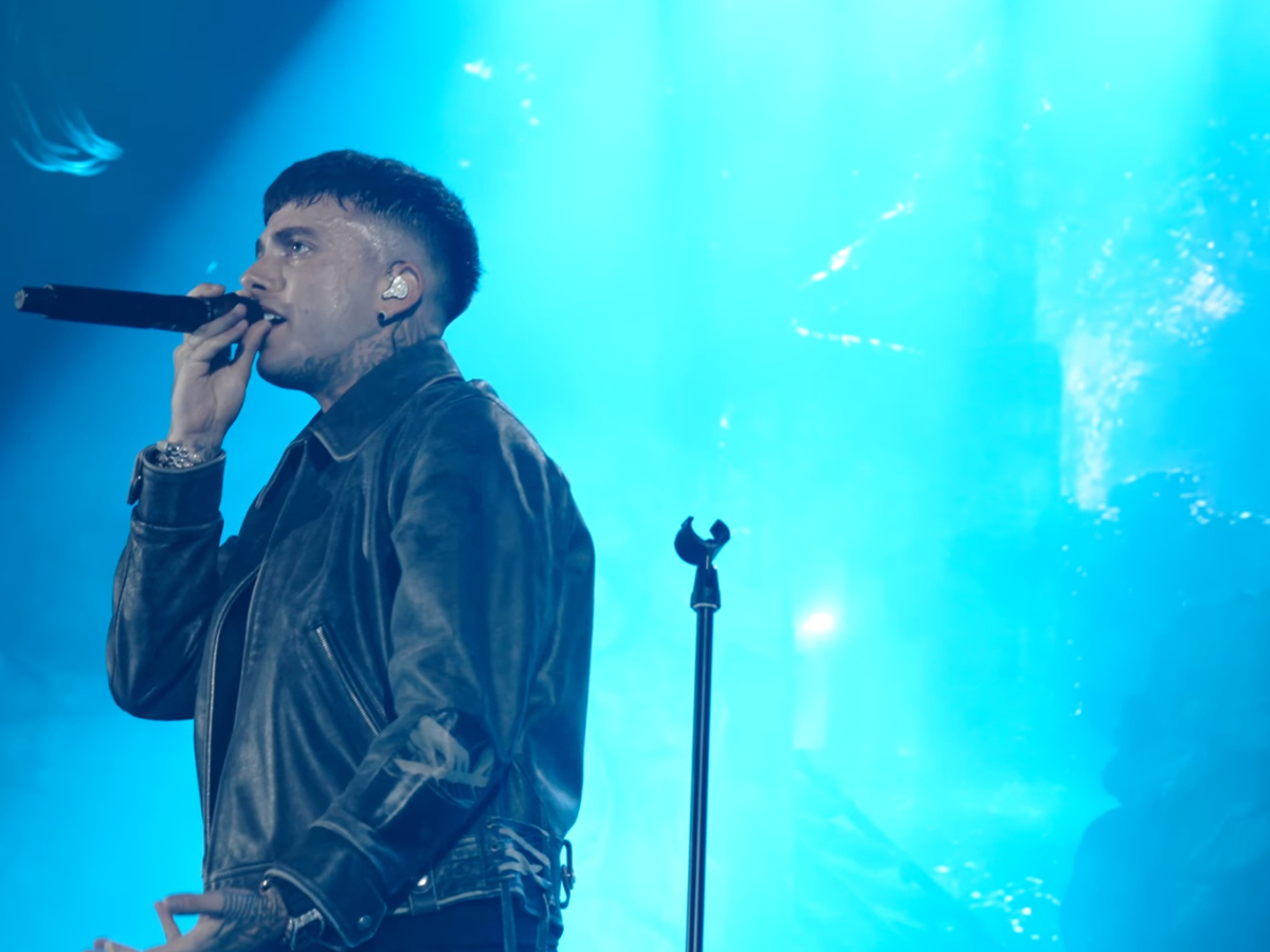 Male singer performing on stage, wearing a black leather jacket and in-ear monitor, holding a microphone under blue stage lights.