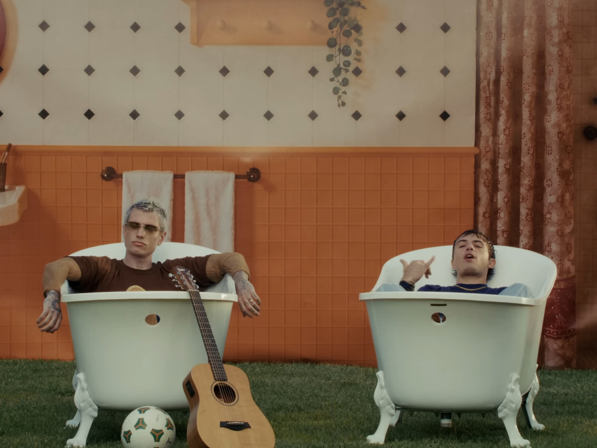 Two men lounging in white clawfoot bathtubs outdoors on grass, with a guitar and a soccer ball nearby against an orange-tiled wall.