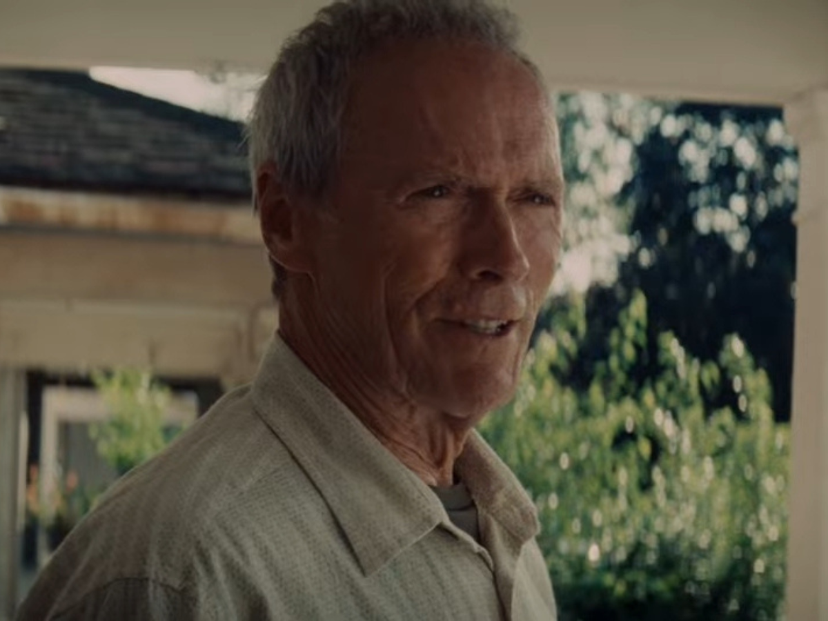 Older man with short gray hair wearing a light shirt, standing on a porch with greenery behind him