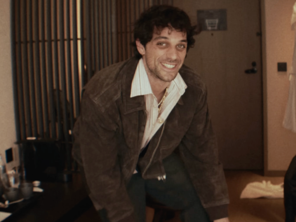 Smiling man in a brown suede jacket leaning forward indoors, wearing a striped shirt and necklace.