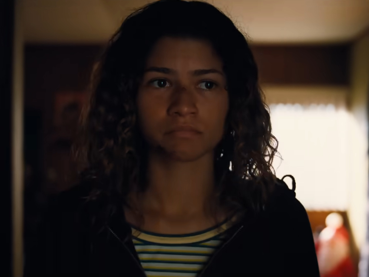 Young woman with curly hair wearing a striped shirt and dark jacket, standing in a dimly lit room with a serious expression.