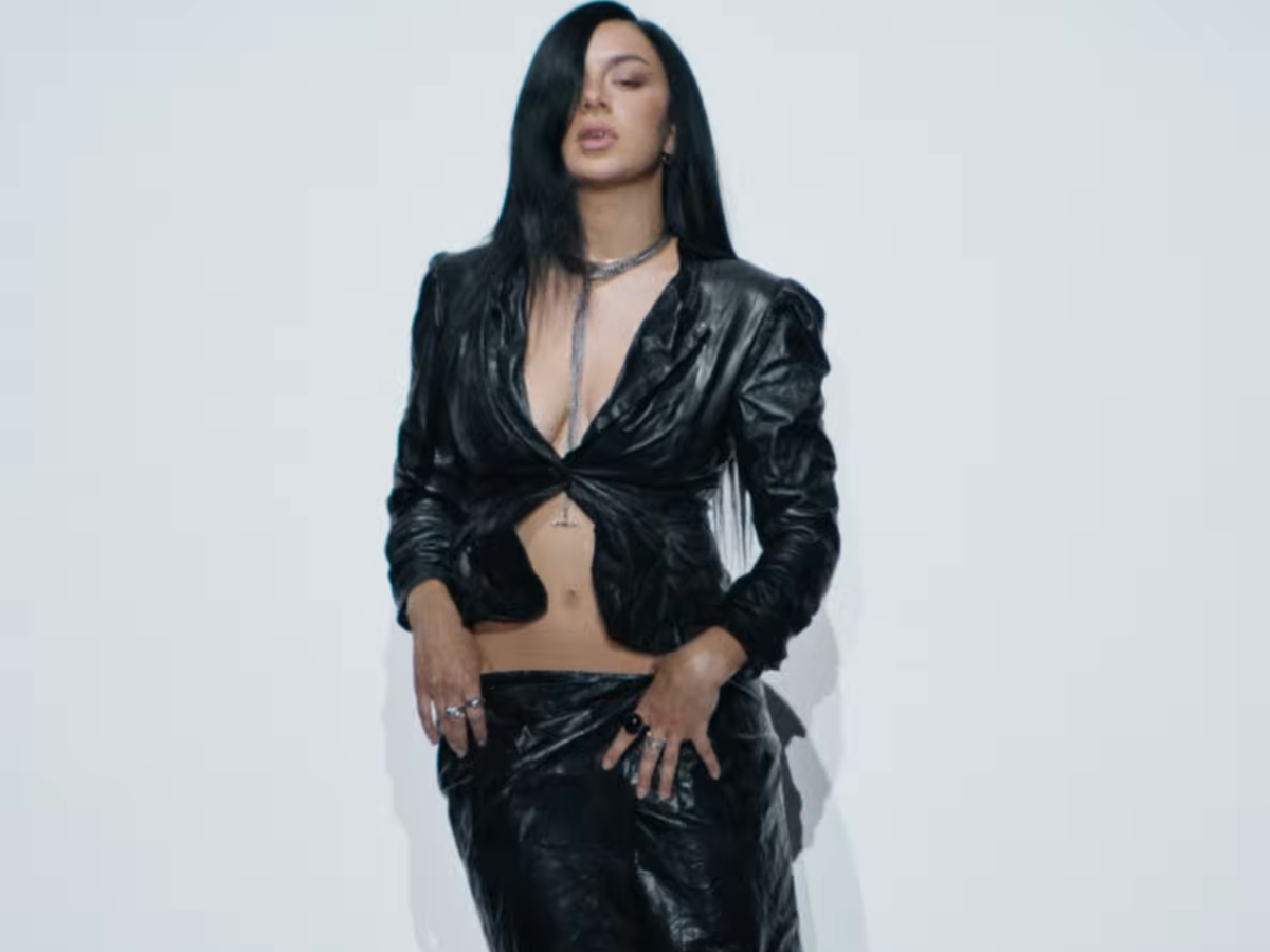 Woman with long black hair wearing a black leather cropped jacket and matching pants, posing with hands on her hips against a pale background.