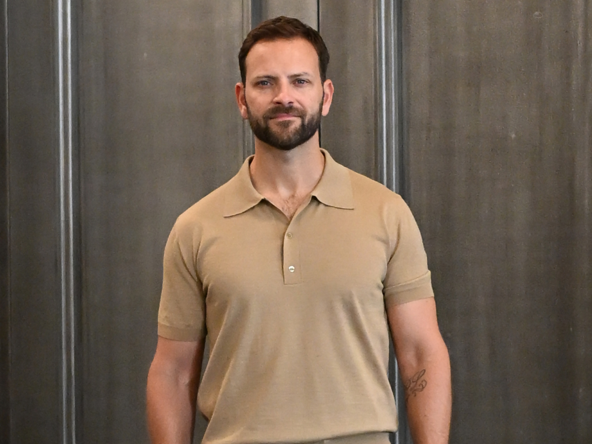 Man with short dark hair and beard stands against a dark panel backdrop, wearing a beige polo shirt.