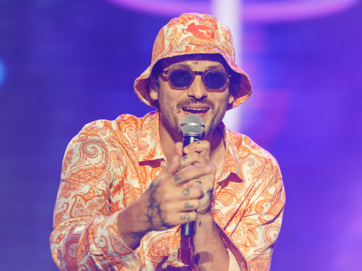Male singer wearing an orange paisley shirt and bucket hat, singing into a microphone on stage with purple lighting in the background.