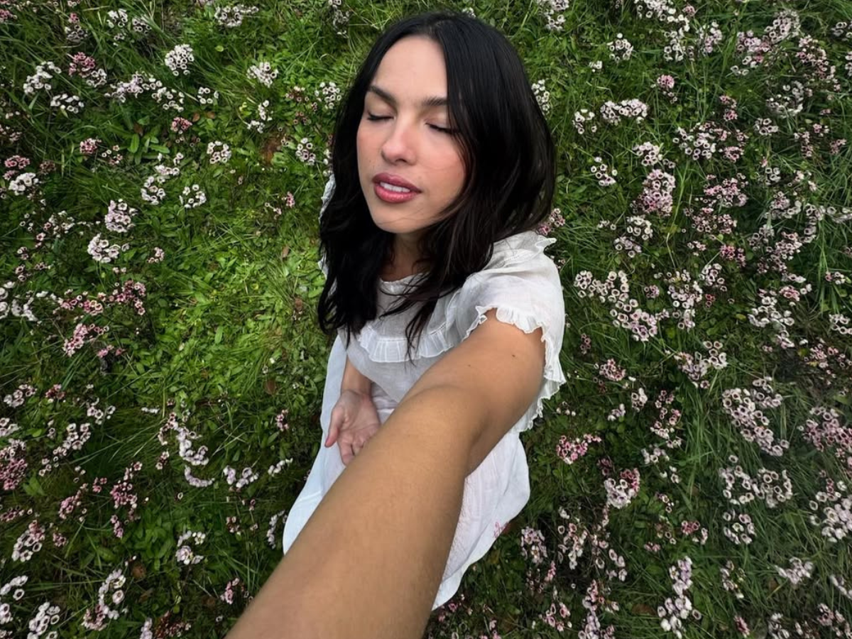 Woman in a white dress takes a selfie with eyes closed in a field of small pink and white flowers.