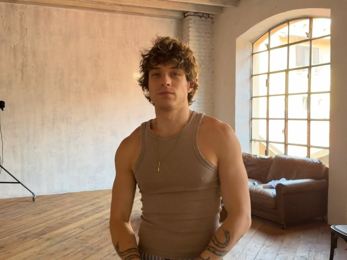 Young man in a brown tank top stands in a sunlit studio with a wooden floor and a large arched window in the background.