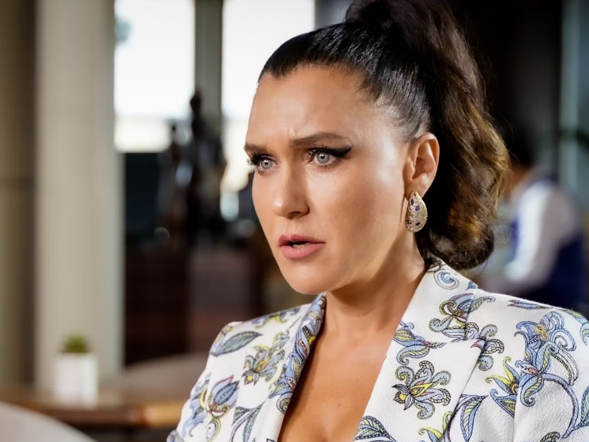 Close-up of a woman with a high ponytail, wearing a patterned white blazer and teardrop earrings, looking to the side.