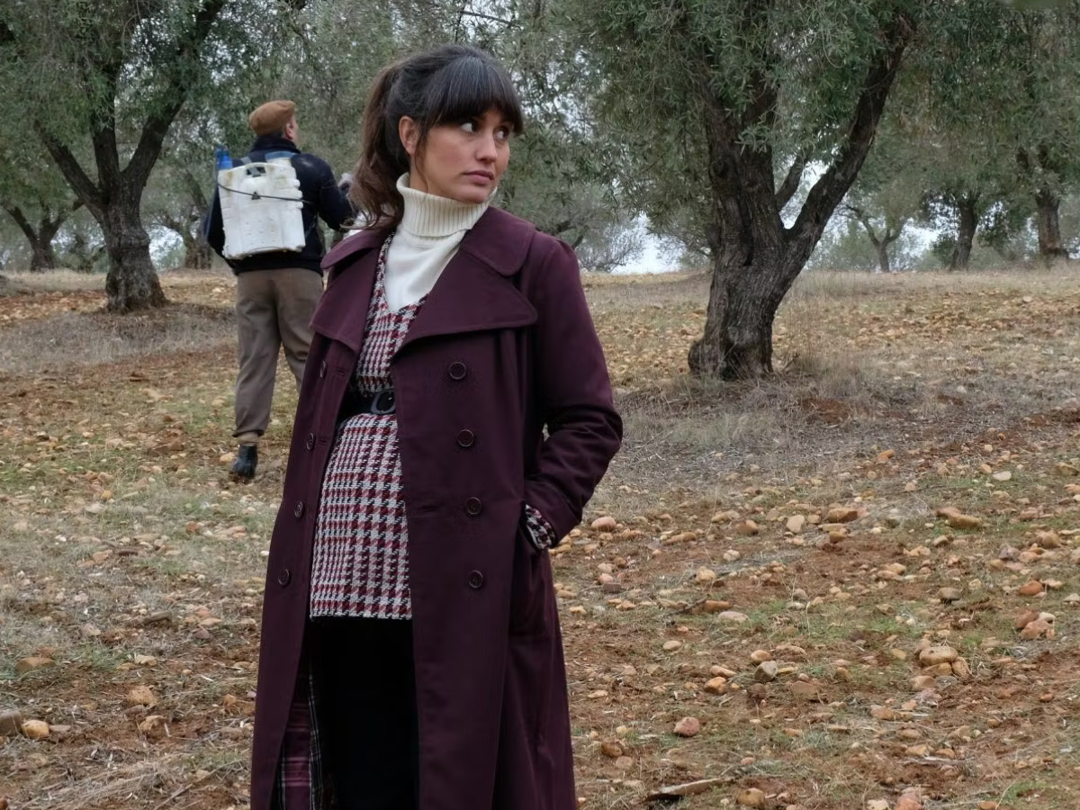 Woman in a burgundy coat stands among olive trees; a man with a backpack sprayer is in the background.