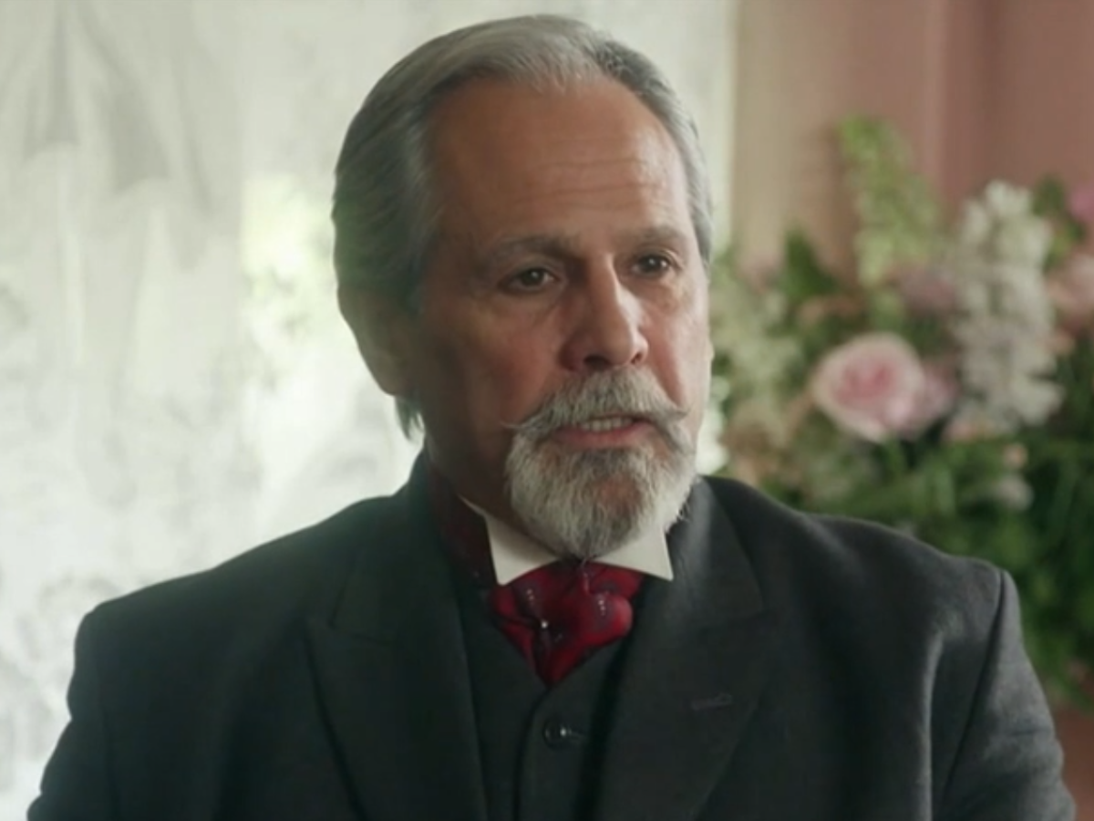 Portrait of an older man with gray hair and a gray beard, wearing a dark suit and red bow tie, indoors with flowers in the background.