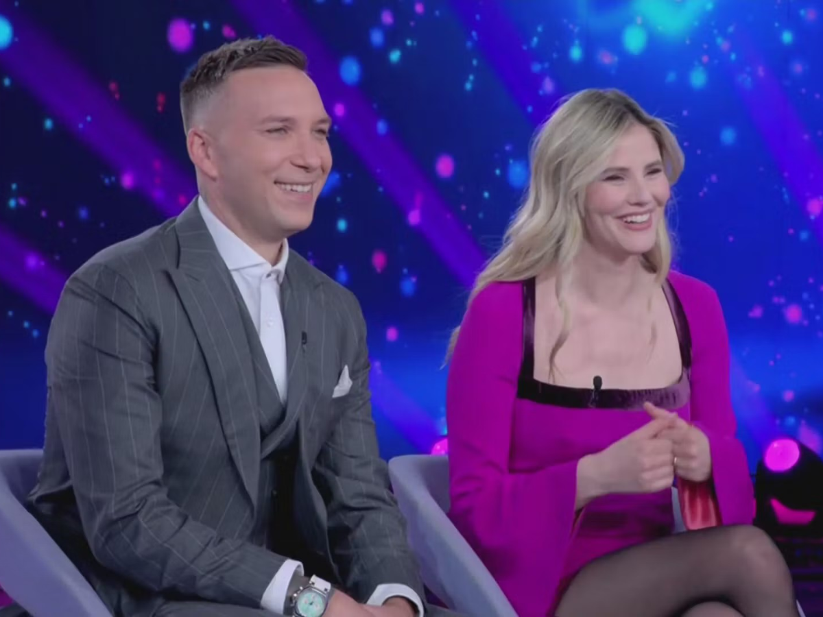 Two smiling TV guests sit on a stage with blue and purple lights in the background.