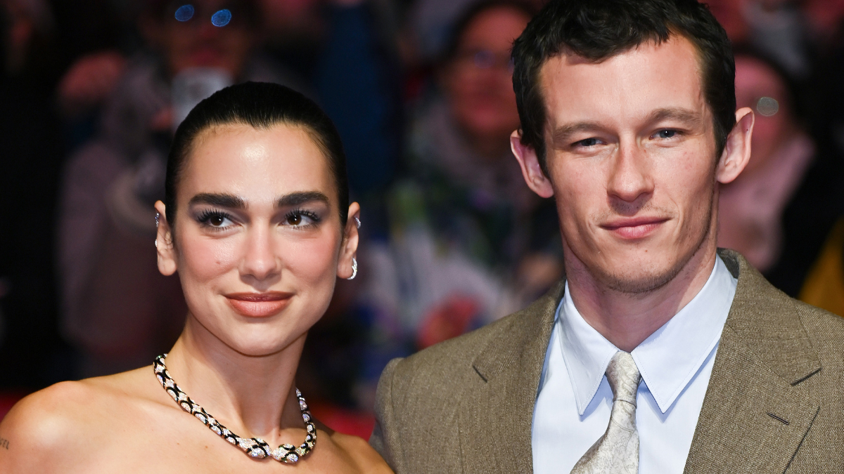 A couple in formal attire posing on the red carpet, woman wearing a necklace and updo, man in a tan suit with a light tie.