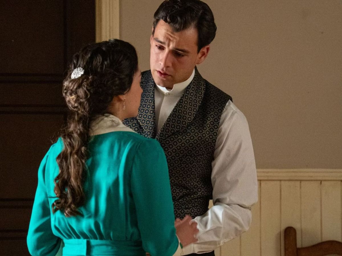 Man in a patterned waistcoat and a woman in a teal dress stand close, gazing at each other in a dimly lit room.