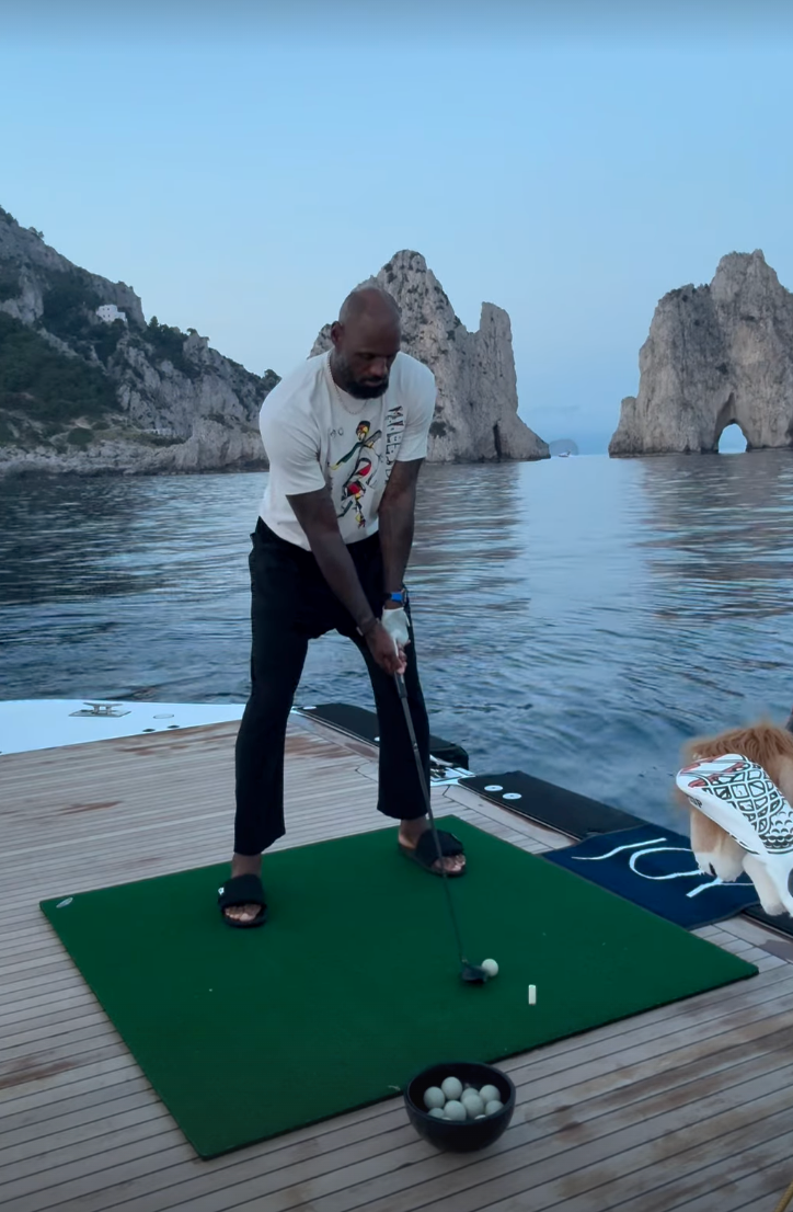 A man on a green mat on a wooden dock by the water, preparing to hit a golf ball with a putter, with a bowl of balls nearby and a dog watching nearby, rocky sea arches in the background.