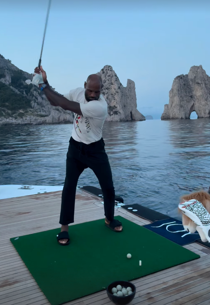 Man swinging a golf club on a boat deck, practicing on a green mat with rocky sea arches in the background and a dog nearby.
