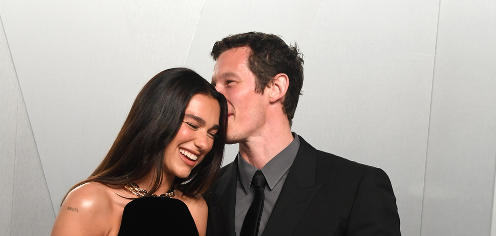 Smiling woman in a black dress stands beside a man in a suit as he gently kisses her on the temple at a formal event.