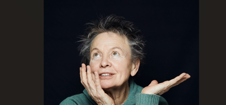 Older woman with short gray hair, green sweater, looking upward with a thoughtful, playful expression and hands near her face.