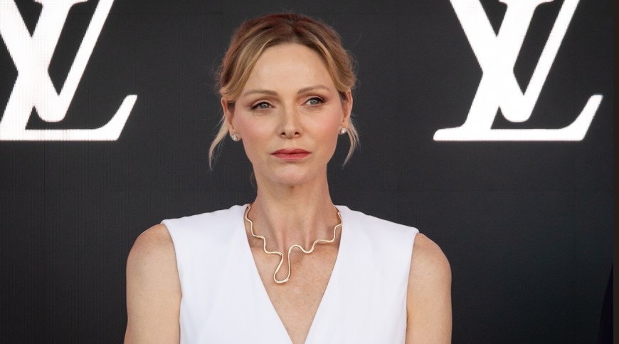 Woman in a white sleeveless dress, wearing a wavy gold necklace, at a fashion event with large LV logos behind her