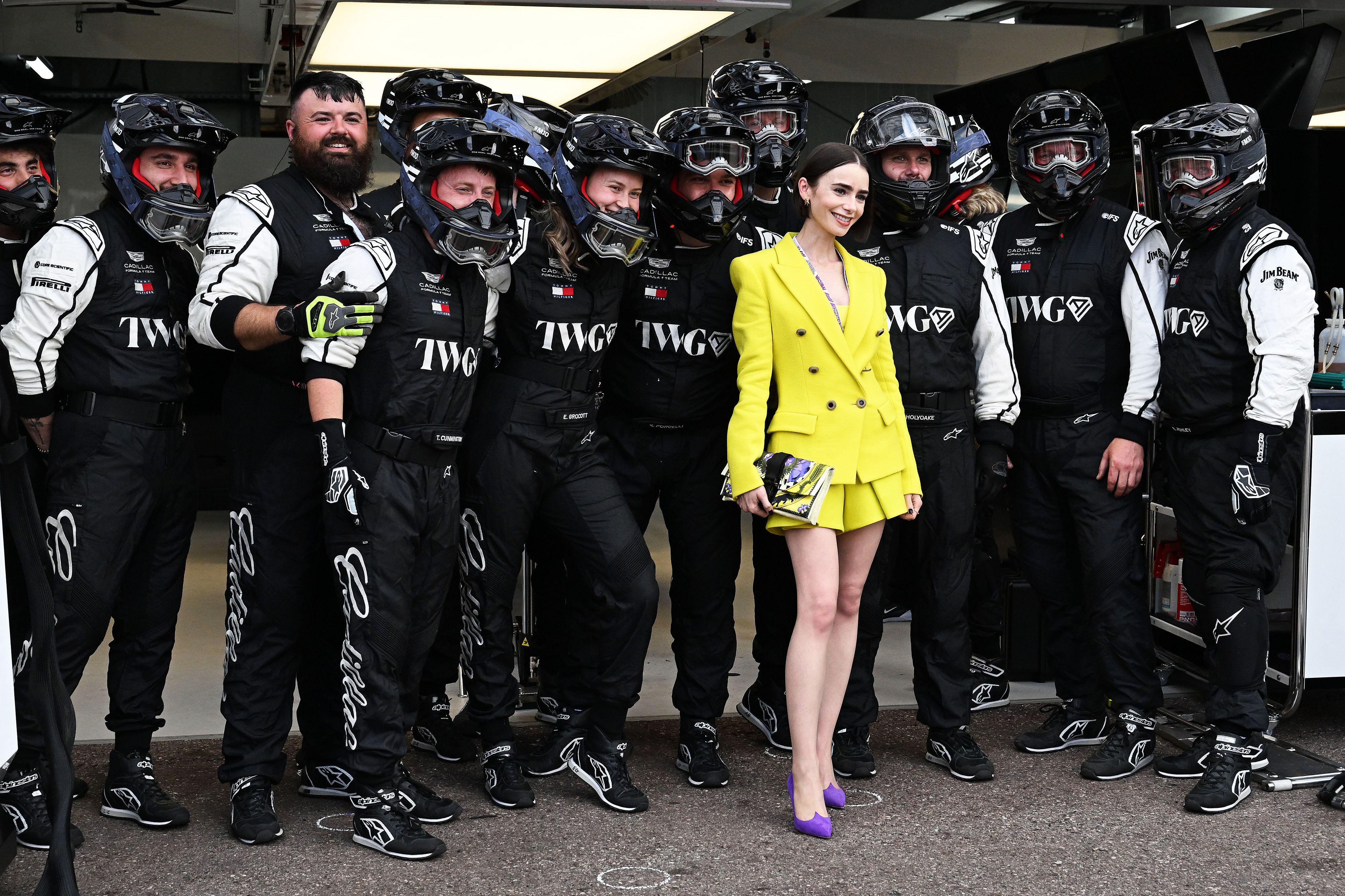 Group of race car drivers in matching black-and-white suits pose with a woman in a bright yellow blazer and purple heels in a garage or pit area.