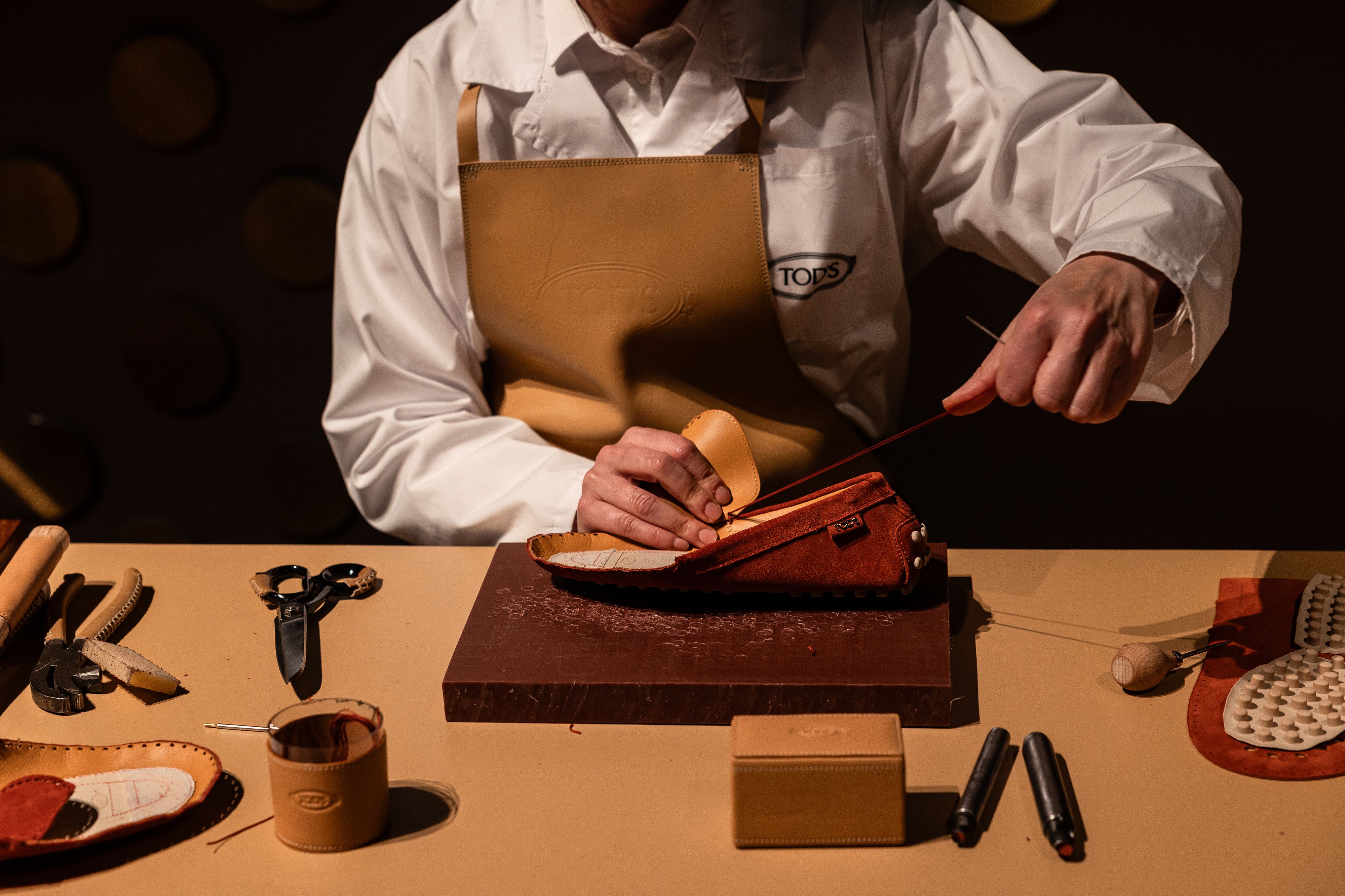 Cobbler sewing a tan leather shoe on a wooden workbench with leather scraps and tools nearby.