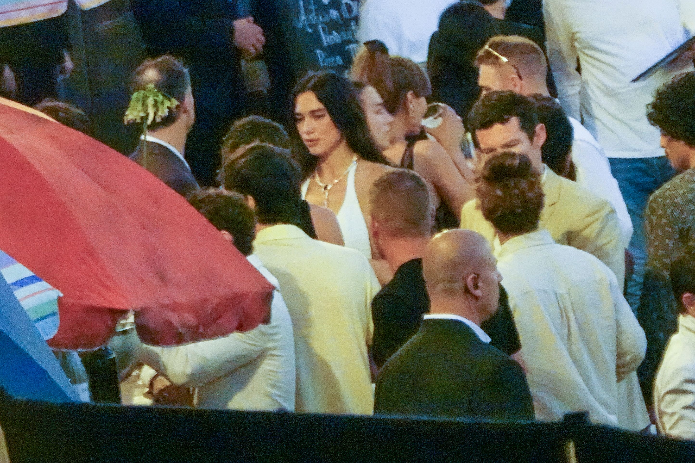 Crowded party scene with people mingling; central figure is a woman in a white dress with a necklace among guests.