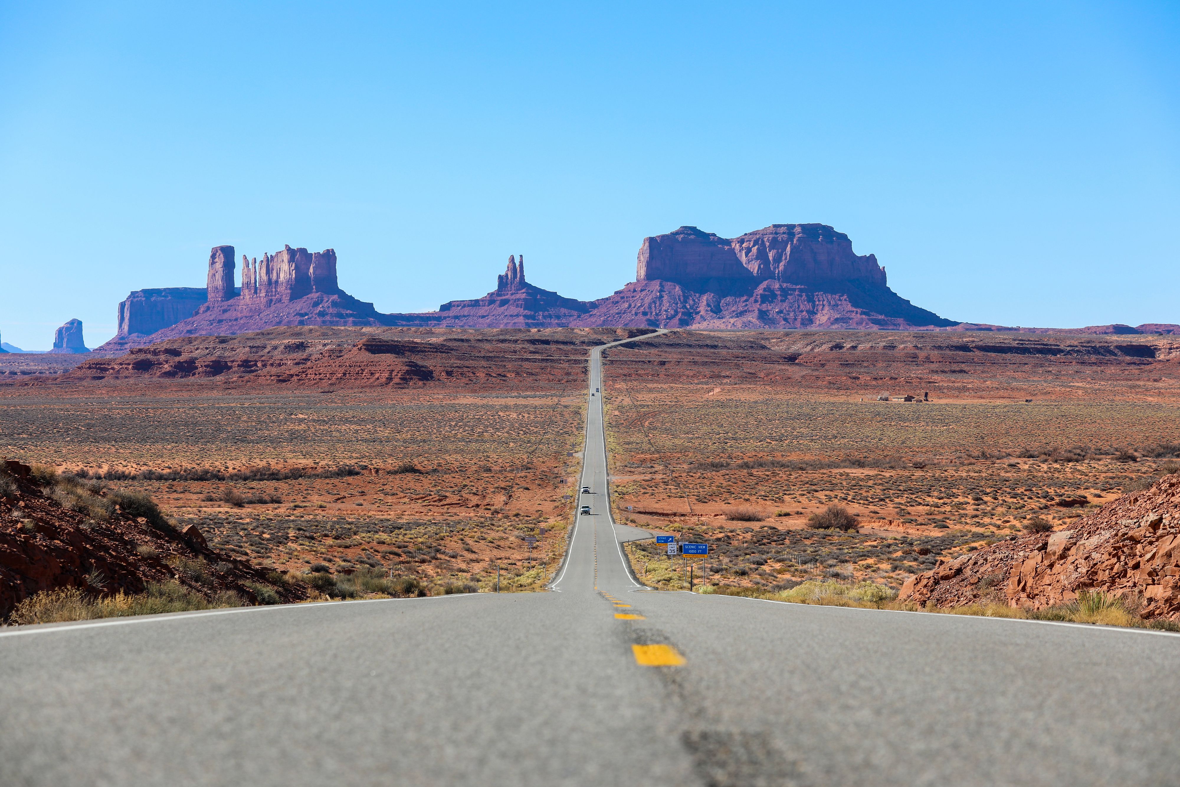 Long straight highway through a desert landscape toward distant red sandstone buttes under a clear blue sky.
