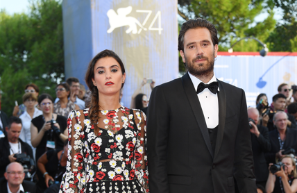 Man in a black tuxedo and a woman in a floral long-sleeve dress pose side by side on a red carpet, with photographers and festival banners in the background.