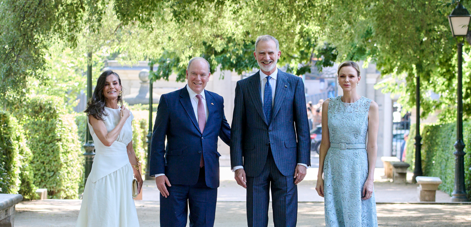 Four well-dressed adults walk down a sunlit garden path with trees and lampposts.