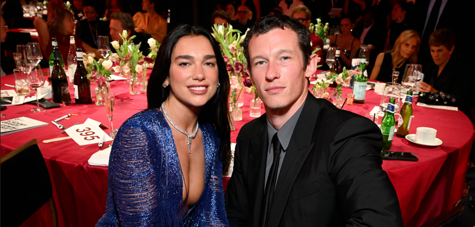 Couple in formal attire seated at a round table with pink tablecloths and floral centerpiece at a gala event, smiling at the camera.