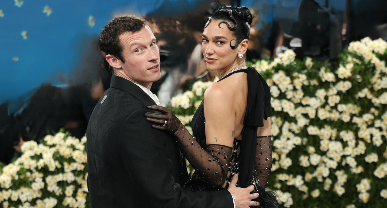 Couple posing on a flower-decorated backdrop at a formal event; man in a tuxedo, woman in a black beaded gown and gloves, looking toward the camera.