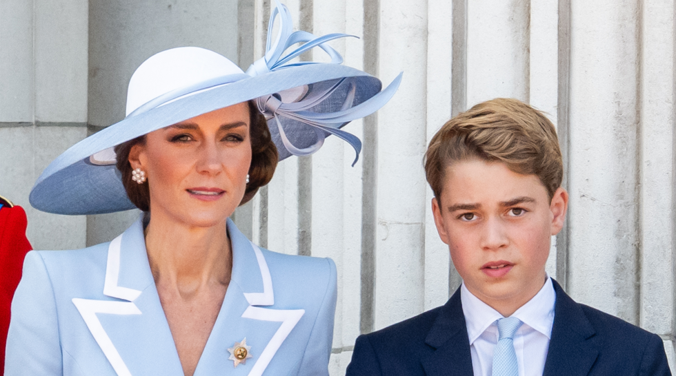 Woman in a pale blue suit and wide hat stands beside a boy in a navy suit against a stone wall.