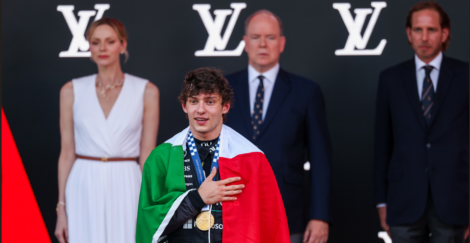 Young athlete with a gold medal draped in the Italian flag, hand over chest, on stage with officials in the background and LV logos behind them.