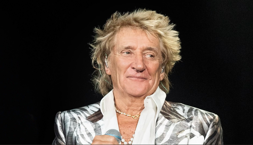 Male singer on stage wearing a silver zebra-pattern blazer and white shirt, holding a microphone with a subtle smile.